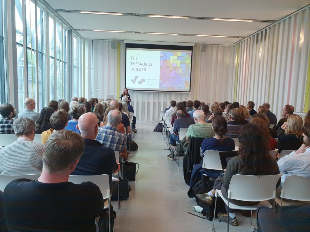 een zaal vol mensen van achterin gezien kijken naar een presentatie over The Thesaurus Builder